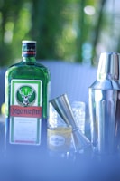 A green bottle with a distinctive label sits beside a silver jigger and a shiny cocktail shaker. A shot glass with the inscription 'Bottoms up' is visible next to the bottle. The background features a soft, blurred greenery.