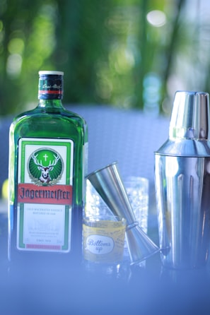A green bottle with a distinctive label sits beside a silver jigger and a shiny cocktail shaker. A shot glass with the inscription 'Bottoms up' is visible next to the bottle. The background features a soft, blurred greenery.