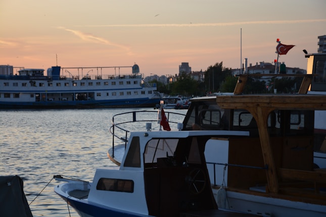 Several boats are docked along a waterfront during sunset, with one prominently displaying a flag. The sky is a mix of warm colors as the sun sets, and the water reflects these hues. In the background, a larger ferry is visible, and a cityscape can be seen with various buildings and trees.
