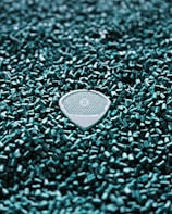 A textured guitar pick is nestled within a field of small turquoise plastic pellets. The pick features an intricate geometric pattern and the letters 'R O B O' at its center.