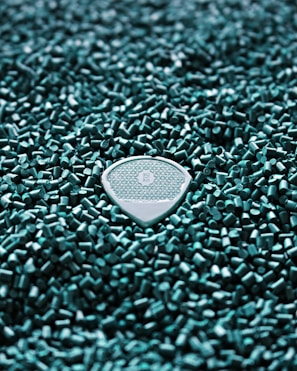 A textured guitar pick is nestled within a field of small turquoise plastic pellets. The pick features an intricate geometric pattern and the letters 'R O B O' at its center.