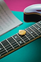 A close-up of an electric guitar neck with a yellow guitar pick resting on the strings. The background features bright colors, including teal and pink, and a blurred sheet of music placed to the side.