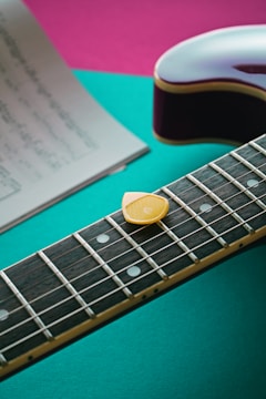 A close-up of an electric guitar neck with a yellow guitar pick resting on the strings. The background features bright colors, including teal and pink, and a blurred sheet of music placed to the side.