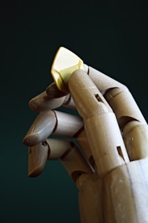 A wooden artist's model hand is holding a small yellow gem between its fingers against a dark green background.