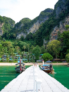 A pier extends into a turquoise sea, bordered by lush greenery and tropical hills. Two traditional long-tail boats are moored by the dock. In the background, trees and hills create a picturesque landscape with resort buildings visible amongst the foliage.