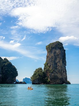 A serene seascape with tall limestone cliffs covered in lush greenery rising prominently from turquoise waters. Two kayakers paddle peacefully in a yellow kayak, exploring the calm sea under a bright blue sky scattered with fluffy white clouds.