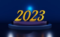 Large golden numbers '2023' are prominently displayed on a circular platform. The background is a deep blue with a central light source casting a blue halo around the numbers.