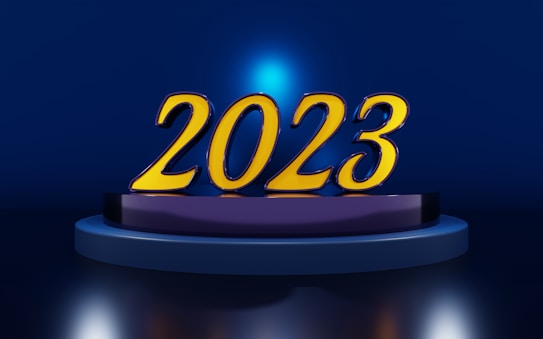 Large golden numbers '2023' are prominently displayed on a circular platform. The background is a deep blue with a central light source casting a blue halo around the numbers.