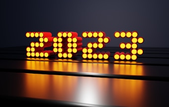 A series of illuminated circular lights form the numbers 2023 against a dark background. The lights are predominantly orange and are arranged in rows to create the numeric shape. The surface they rest on is reflective, giving a glossy appearance, and enhancing the glow of the lights.