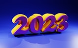 Bright and colorful 3D numbers displaying '2023' are prominently positioned in the center. The numbers are stylized with a vibrant purple and yellow color scheme, casting shadows on a blue background. The lighting creates a glossy and reflective effect on the numbers.