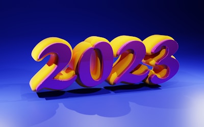Bright and colorful 3D numbers displaying '2023' are prominently positioned in the center. The numbers are stylized with a vibrant purple and yellow color scheme, casting shadows on a blue background. The lighting creates a glossy and reflective effect on the numbers.