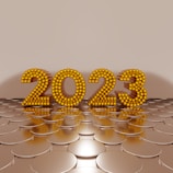 Golden numbers forming '2023' are made of spherical elements and stand upright on a reflective, circular pattern surface, set against a plain beige background.
