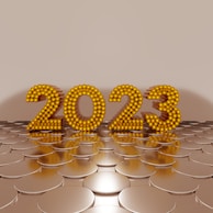Golden numbers forming '2023' are made of spherical elements and stand upright on a reflective, circular pattern surface, set against a plain beige background.