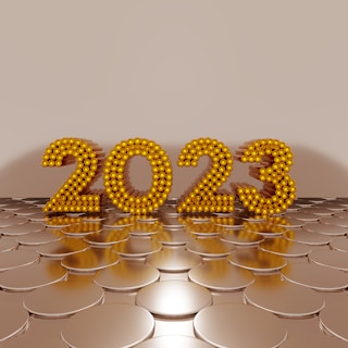 Golden numbers forming '2023' are made of spherical elements and stand upright on a reflective, circular pattern surface, set against a plain beige background.