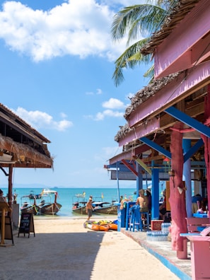 A sunny beach scene with long-tail boats docked near the shore. Colorful beach bars with rustic wooden frames and thatched roofs line the path leading to the water. People are sitting and relaxing both inside and outside the bars, enjoying the tropical atmosphere. There is a clear blue sky with scattered white clouds overhead, and a few palm fronds hang into the frame.