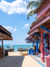 A sunny beach scene with long-tail boats docked near the shore. Colorful beach bars with rustic wooden frames and thatched roofs line the path leading to the water. People are sitting and relaxing both inside and outside the bars, enjoying the tropical atmosphere. There is a clear blue sky with scattered white clouds overhead, and a few palm fronds hang into the frame.
