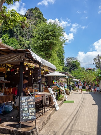 A street scene with market stalls on the left side, offering BBQ menu items such as red snapper and tuna fish, displayed on a chalkboard. A vendor is grilling food on an open barbecue. Wooden tables and chairs are set up inside the market area, while a tree and lush greenery in the background indicate an outdoor setting. Several people are walking along the paved path on a sunny day.