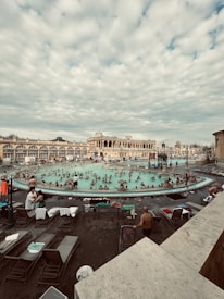 A bustling outdoor thermal bath area surrounded by historical architecture. Numerous people are enjoying the warm water, with lounge chairs and towels scattered around the poolside. The sky is filled with a layer of soft clouds, adding a serene backdrop to the lively scene.