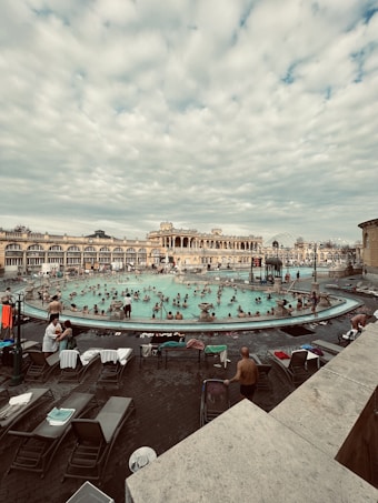 A bustling outdoor thermal bath area surrounded by historical architecture. Numerous people are enjoying the warm water, with lounge chairs and towels scattered around the poolside. The sky is filled with a layer of soft clouds, adding a serene backdrop to the lively scene.