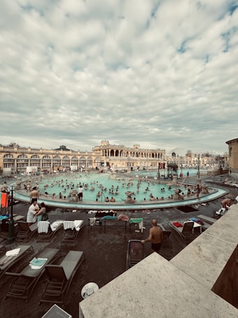 A bustling outdoor thermal bath area surrounded by historical architecture. Numerous people are enjoying the warm water, with lounge chairs and towels scattered around the poolside. The sky is filled with a layer of soft clouds, adding a serene backdrop to the lively scene.