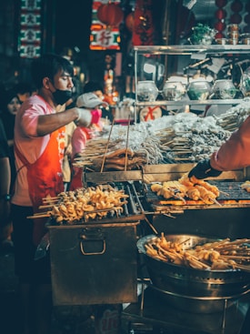 A street vendor is actively grilling skewers of food over a metal grill in a bustling night market. The vendor is wearing a red apron and a black face mask. Various ingredients are on display, including skewers of seafood or meat, possibly surrounded by containers with additional items. Colorful lanterns and vibrant signs, typical of an Asian street market, hang in the background, adding to the lively atmosphere.
