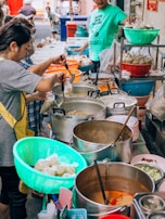 Several people are preparing and serving food at an outdoor market stall. Pots filled with various soups or stews are being stirred, and plastic baskets hold ingredients or packaged items. The atmosphere is busy and lively, with containers and utensils scattered around the workspace.
