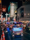 A bustling nighttime street scene featuring vibrant tuk-tuks illuminated by colorful lights, lined up along a busy road. The street is crowded with pedestrians and motorcycles, with numerous neon signs and advertisements glowing on the buildings. A prominent hotel sign can be seen in the background.
