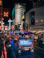 A bustling nighttime street scene featuring vibrant tuk-tuks illuminated by colorful lights, lined up along a busy road. The street is crowded with pedestrians and motorcycles, with numerous neon signs and advertisements glowing on the buildings. A prominent hotel sign can be seen in the background.