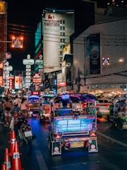 A bustling nighttime street scene featuring vibrant tuk-tuks illuminated by colorful lights, lined up along a busy road. The street is crowded with pedestrians and motorcycles, with numerous neon signs and advertisements glowing on the buildings. A prominent hotel sign can be seen in the background.