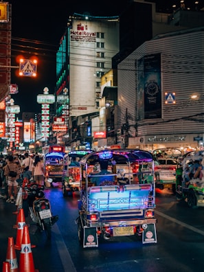 A bustling nighttime street scene featuring vibrant tuk-tuks illuminated by colorful lights, lined up along a busy road. The street is crowded with pedestrians and motorcycles, with numerous neon signs and advertisements glowing on the buildings. A prominent hotel sign can be seen in the background.