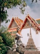 A woman poses stylishly in front of an ornate temple with intricate details and vibrant colors. She stands on a stone sculpture, surrounded by lush green foliage. The temple features golden accents and a distinct architectural style.