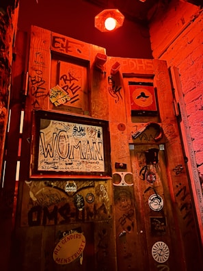 A dimly lit space features a wooden door covered in a variety of graffiti and stickers. There's a rectangular frame with the word 'WOMAN' prominently displayed. The lighting casts a reddish-orange hue over the scene, creating a shadowy atmosphere. Several stickers and scribbles cover the surface, contributing to a gritty, urban aesthetic. Above, there's a glowing red light fixture.