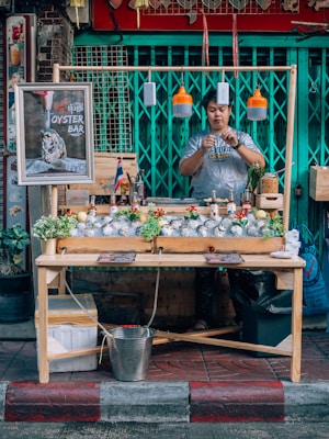 A street vendor operates an oyster bar stall, featuring a wooden table adorned with fresh oysters and plants. The vendor, wearing a casual t-shirt, prepares food behind the table. Hanging above are modern pendant lights, while various condiments and decorations, including a small flag, are displayed on the table. The background consists of a closed metal gate painted teal.