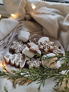 A cozy scene featuring a plate of decorated gingerbread cookies in the shapes of snowflakes and trees, accompanied by marshmallows. The setup is surrounded by a soft beige blanket and lit by warm fairy lights, giving a festive and inviting feel.