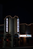 A nighttime view of a theater entrance, illuminated by vertical and horizontal signs displaying 'THEATRE' and other text. The building is adorned with festive string lights, creating a classic, inviting atmosphere. The environment is dimly lit, emphasizing the glow of the theater lights.