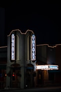 A nighttime view of a theater entrance, illuminated by vertical and horizontal signs displaying 'THEATRE' and other text. The building is adorned with festive string lights, creating a classic, inviting atmosphere. The environment is dimly lit, emphasizing the glow of the theater lights.