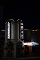 A nighttime view of a theater entrance, illuminated by vertical and horizontal signs displaying 'THEATRE' and other text. The building is adorned with festive string lights, creating a classic, inviting atmosphere. The environment is dimly lit, emphasizing the glow of the theater lights.