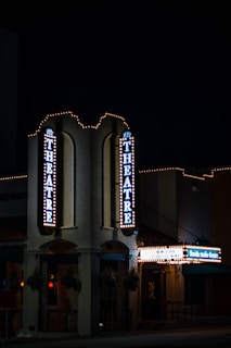 A nighttime view of a theater entrance, illuminated by vertical and horizontal signs displaying 'THEATRE' and other text. The building is adorned with festive string lights, creating a classic, inviting atmosphere. The environment is dimly lit, emphasizing the glow of the theater lights.