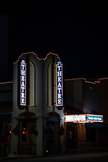 A nighttime view of a theater entrance, illuminated by vertical and horizontal signs displaying 'THEATRE' and other text. The building is adorned with festive string lights, creating a classic, inviting atmosphere. The environment is dimly lit, emphasizing the glow of the theater lights.