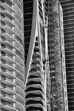 Tall, modern buildings with curved balconies and glass facades dominate the image. A construction scaffold is visible alongside one structure, suggesting ongoing development. The architectural design showcases sleek and futuristic aesthetics.