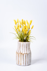 A decorative vase features a rustic design with vertical white and brown streaks. It holds an arrangement of tall, yellow faux flowers with green leaves. The background is a plain, soft white, enhancing the vibrant colors of the flowers.