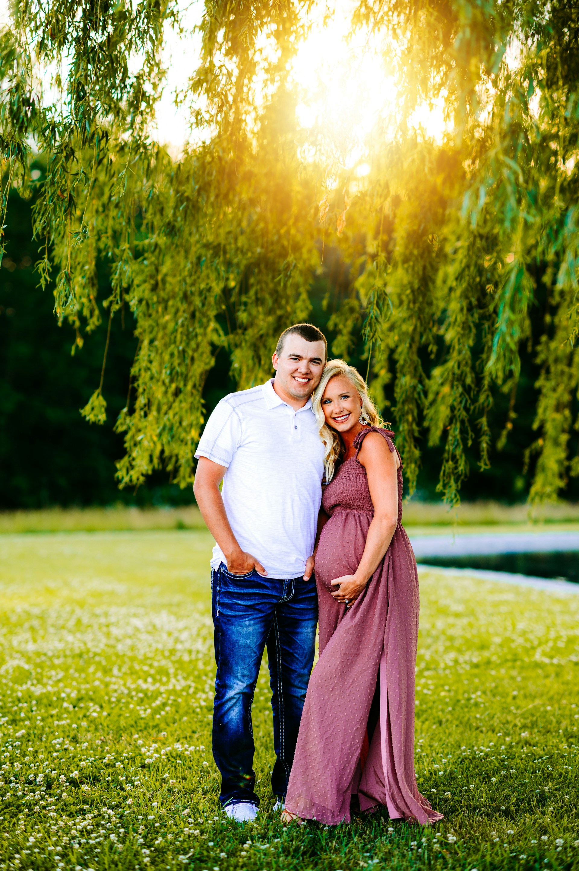 a pregnant couple standing in the grass under a tree
