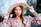 A woman with long brown hair wears a wide-brimmed beige hat and a colorful floral blouse. She holds the brim of her hat, with well-manicured nails painted in a bright color. Her expression is confident, and she is accessorized with hoop earrings and bracelets.