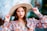A woman with long brown hair wears a wide-brimmed beige hat and a colorful floral blouse. She holds the brim of her hat, with well-manicured nails painted in a bright color. Her expression is confident, and she is accessorized with hoop earrings and bracelets.
