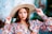 A woman with long brown hair wears a wide-brimmed beige hat and a colorful floral blouse. She holds the brim of her hat, with well-manicured nails painted in a bright color. Her expression is confident, and she is accessorized with hoop earrings and bracelets.