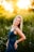 A woman with blonde hair poses in a field of wildflowers under warm, golden sunlight. She is wearing a denim dress and smiling, with one hand resting on her hip. The background is blurred, highlighting the greenery and vibrant floral environment.
