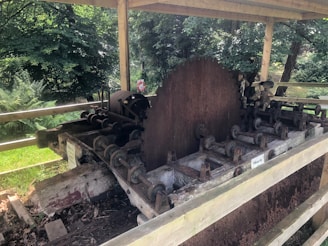 An old, rusted sawmill machine with a large circular saw blade. The machine is situated outdoors under a wooden shelter, surrounded by a lush green forest. The structure shows signs of wear and aging, with several mechanical components and rollers.