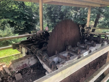 An old, rusted sawmill machine with a large circular saw blade. The machine is situated outdoors under a wooden shelter, surrounded by a lush green forest. The structure shows signs of wear and aging, with several mechanical components and rollers.