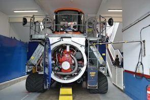A large industrial machine with an open casing revealing complex internal mechanisms, including pipes, wiring, and a prominently visible red component. The machine is indoors, in what appears to be a maintenance or workshop area. There are barriers around the machine with a visible safety warning 'KEEP OFF'. In the background, a staircase and a few people are visible.
