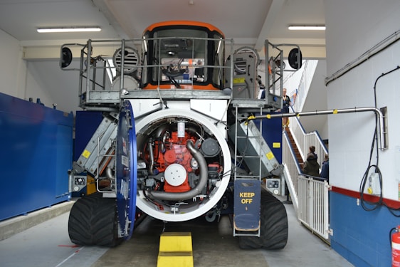 A large industrial machine with an open casing revealing complex internal mechanisms, including pipes, wiring, and a prominently visible red component. The machine is indoors, in what appears to be a maintenance or workshop area. There are barriers around the machine with a visible safety warning 'KEEP OFF'. In the background, a staircase and a few people are visible.